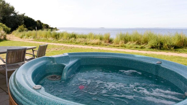 A hot tub with a view of the sea, surrounded by a grassy area and a wooden deck with chairs.