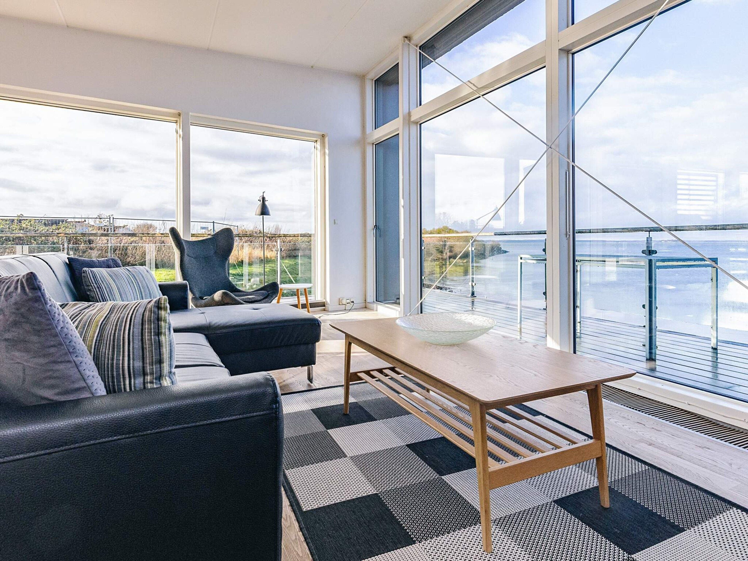 Ein modernes Wohnzimmer mit Sofa, Sessel, Tisch und großen Fenstern mit Blick auf ein Gewässer.
