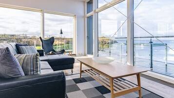 Ein modernes Wohnzimmer mit Sofa, Sessel, Tisch und großen Fenstern mit Blick auf ein Gewässer.