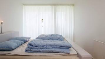 A bedroom with a bed, pillows, a bedside table, a lamp, and a window with curtains.