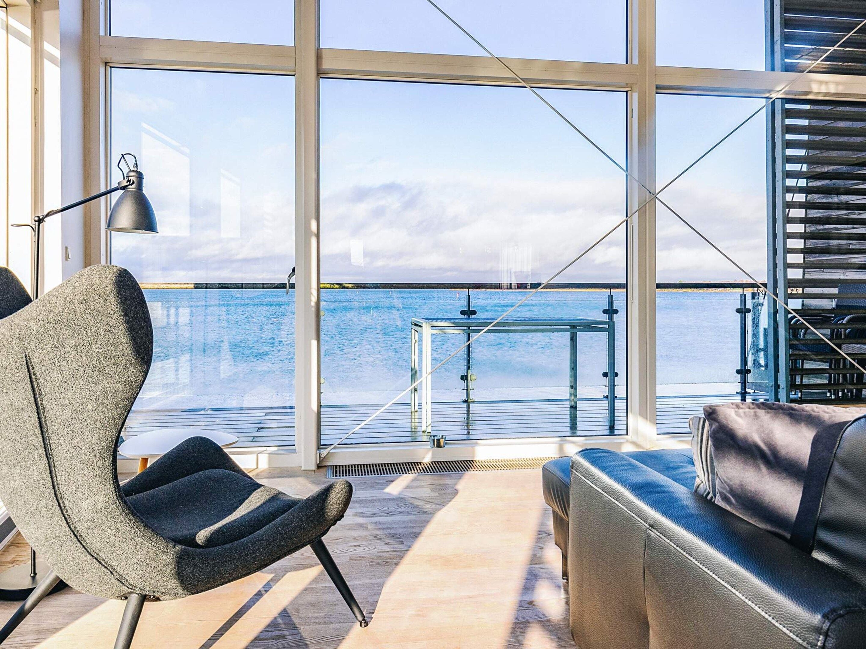 Ein modernes Interieur mit einem großen Fenster mit Blick auf ein Gewässer, eine Sitzecke mit Sofa und Sesseln sowie einem Metalltisch.