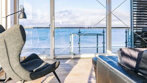 A modern interior with a large window overlooking a body of water, a seating area with a sofa and armchair, and a metal table.