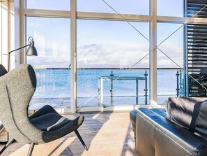 Een modern interieur met een groot raam met uitzicht op het water, een zithoek met een bank en fauteuil, en een metalen tafel.