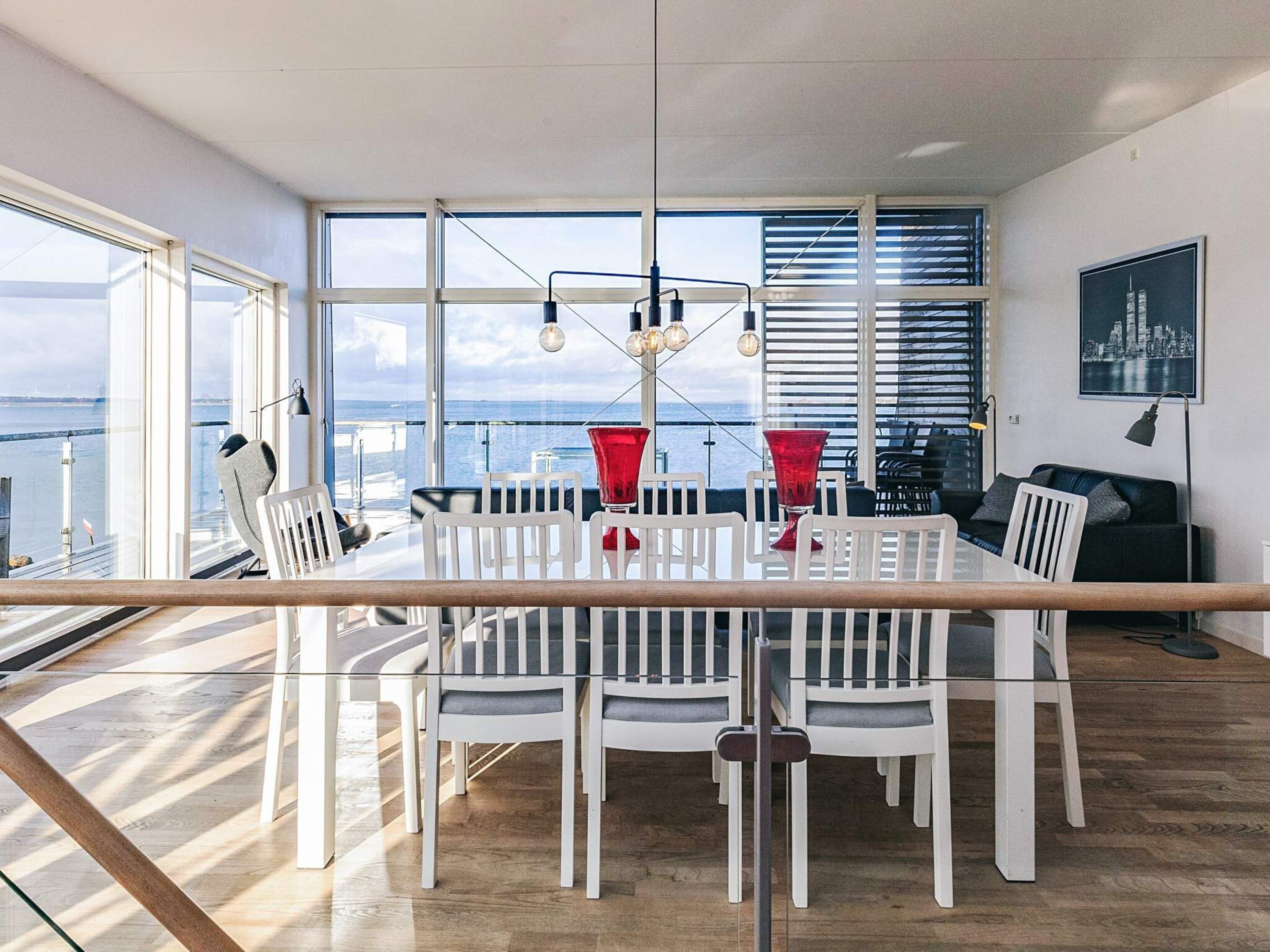 Un comedor moderno con mesa de vidrio y sillas blancas, ventanas amplias con vista a la ciudad, y un cuadro de paisaje urbano en la pared.