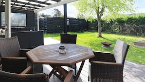 En udendørs patio med et træbord og fletstole, et overdækket område med et metal tag og udsigt til en grøn have med et træ og et hegn.