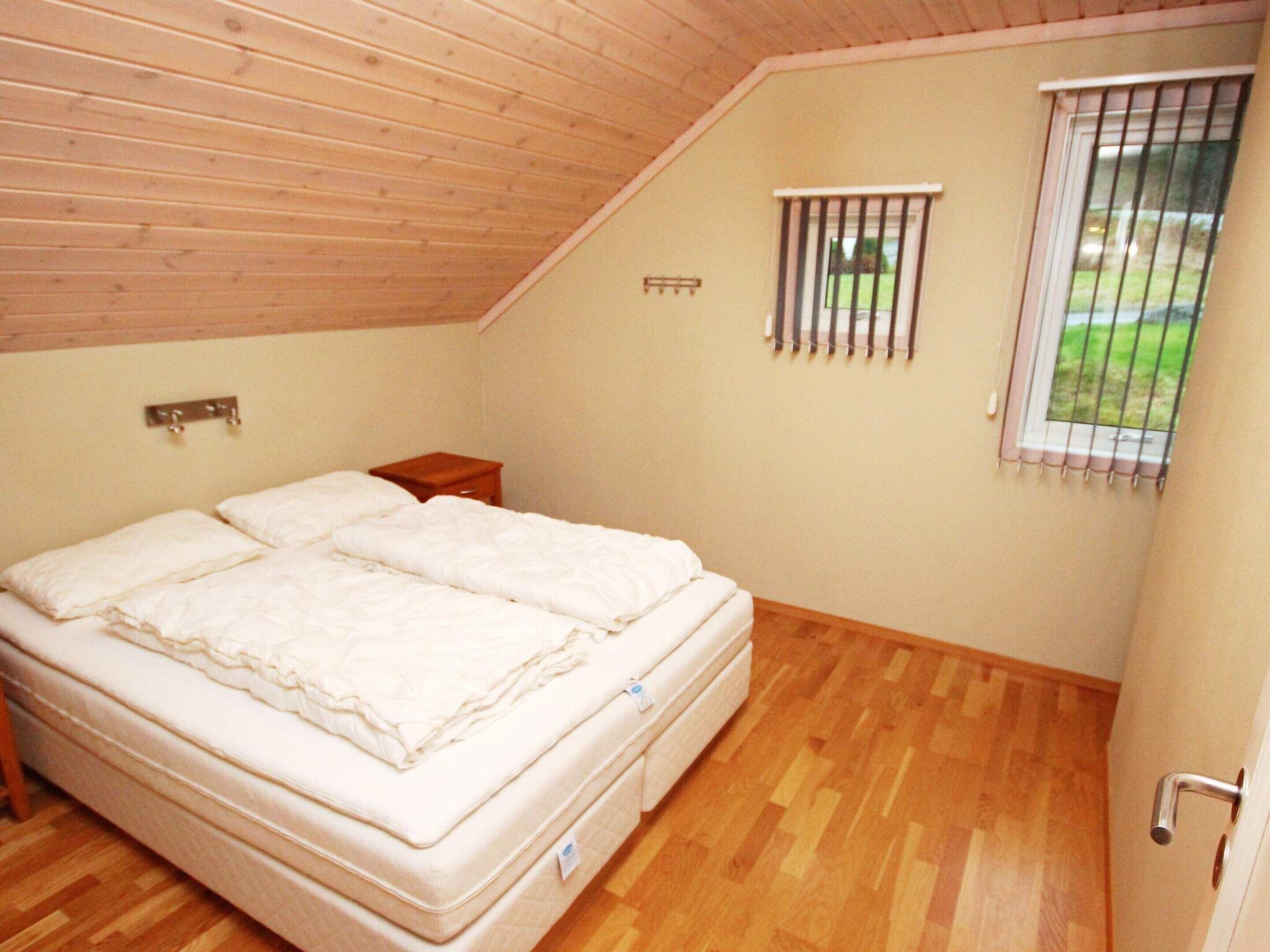A bedroom with a wooden ceiling, a single bed, a small bedside table, a window with bars, and a view of the outdoors.