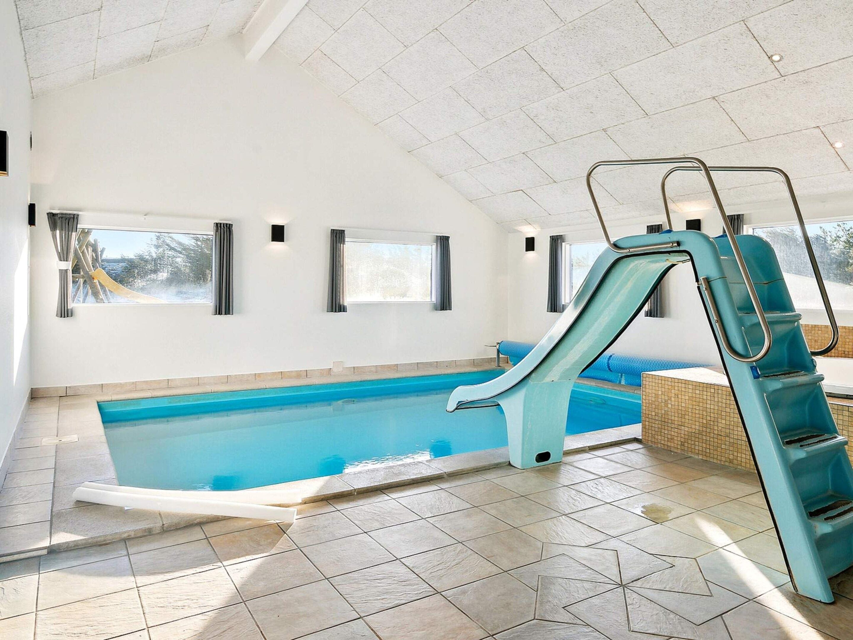 En indendørs swimmingpool med rutsjebane, flisegulv og vinduer.