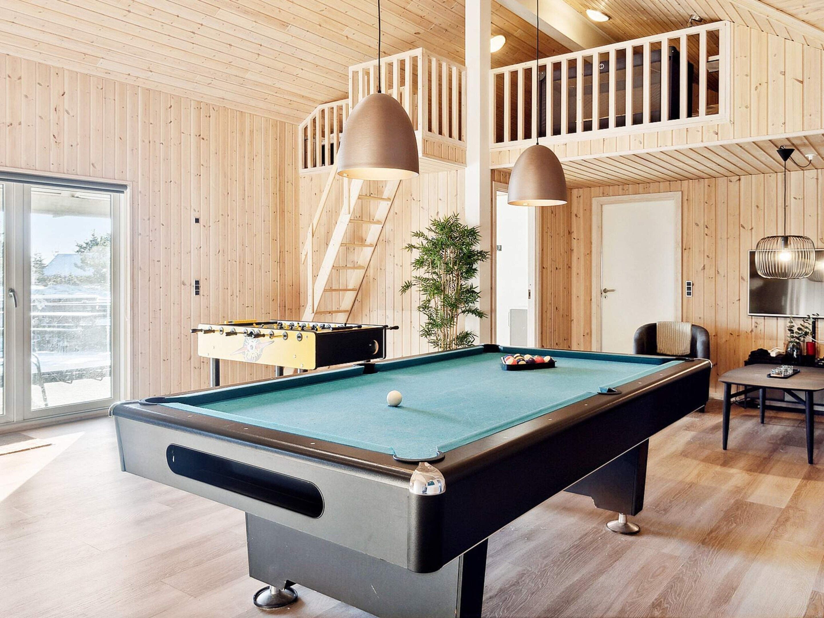 Una mesa de billar junto a una mesa de futbolín y un soporte para tacos en una habitación con paredes de madera y un balcón.