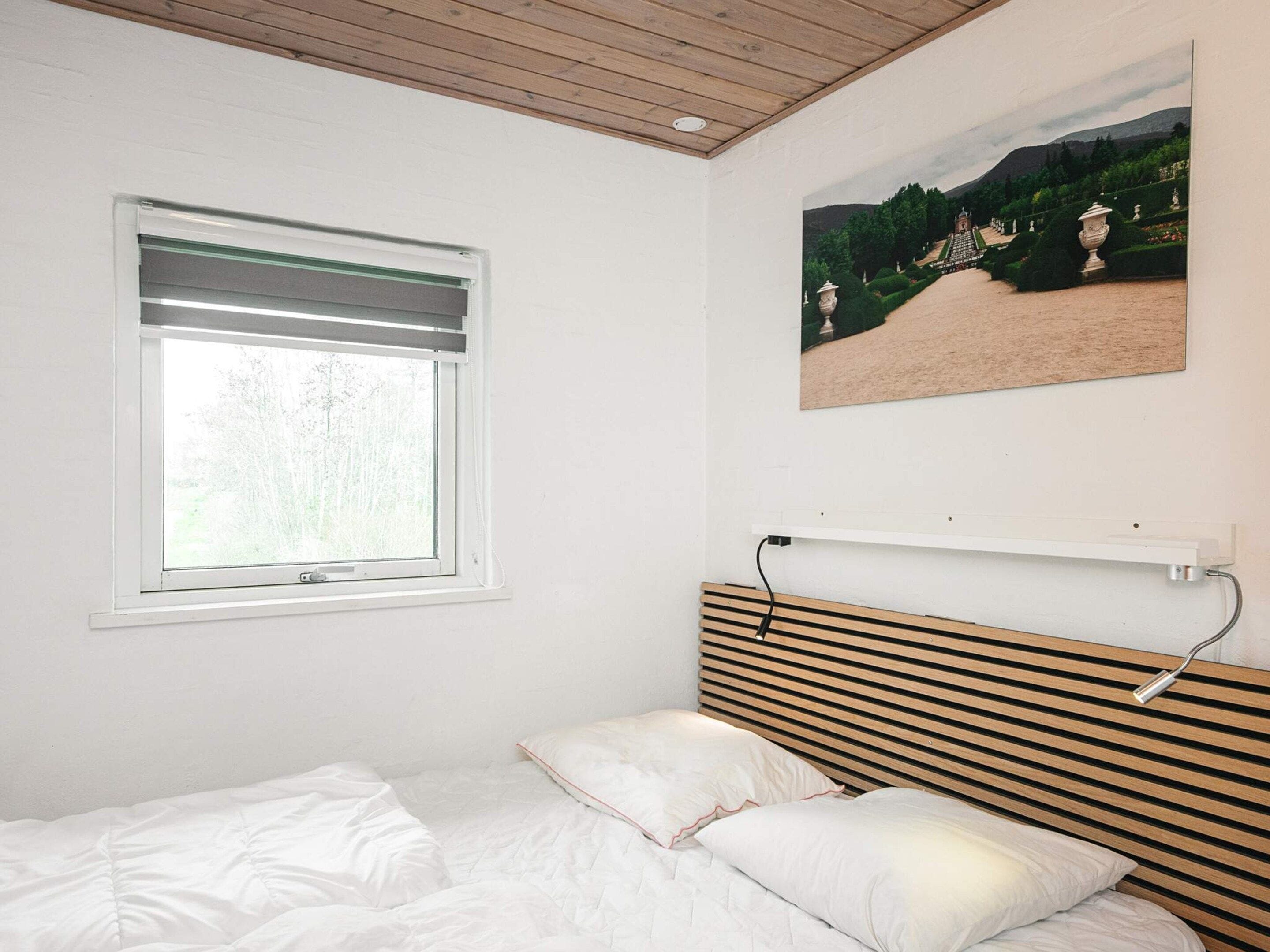 A bedroom with a bed, a window with blinds, a wall-mounted shelf, and a landscape painting.
