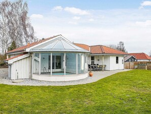 Une piscine extérieure couverte, protégée par une structure en verre, entourée d’une pelouse et bordée d’un bâtiment blanc doté d’un toit en tuiles.