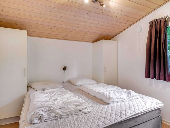 Un dormitorio con cama, techo de madera, paredes blancas y una ventana con cortinas.