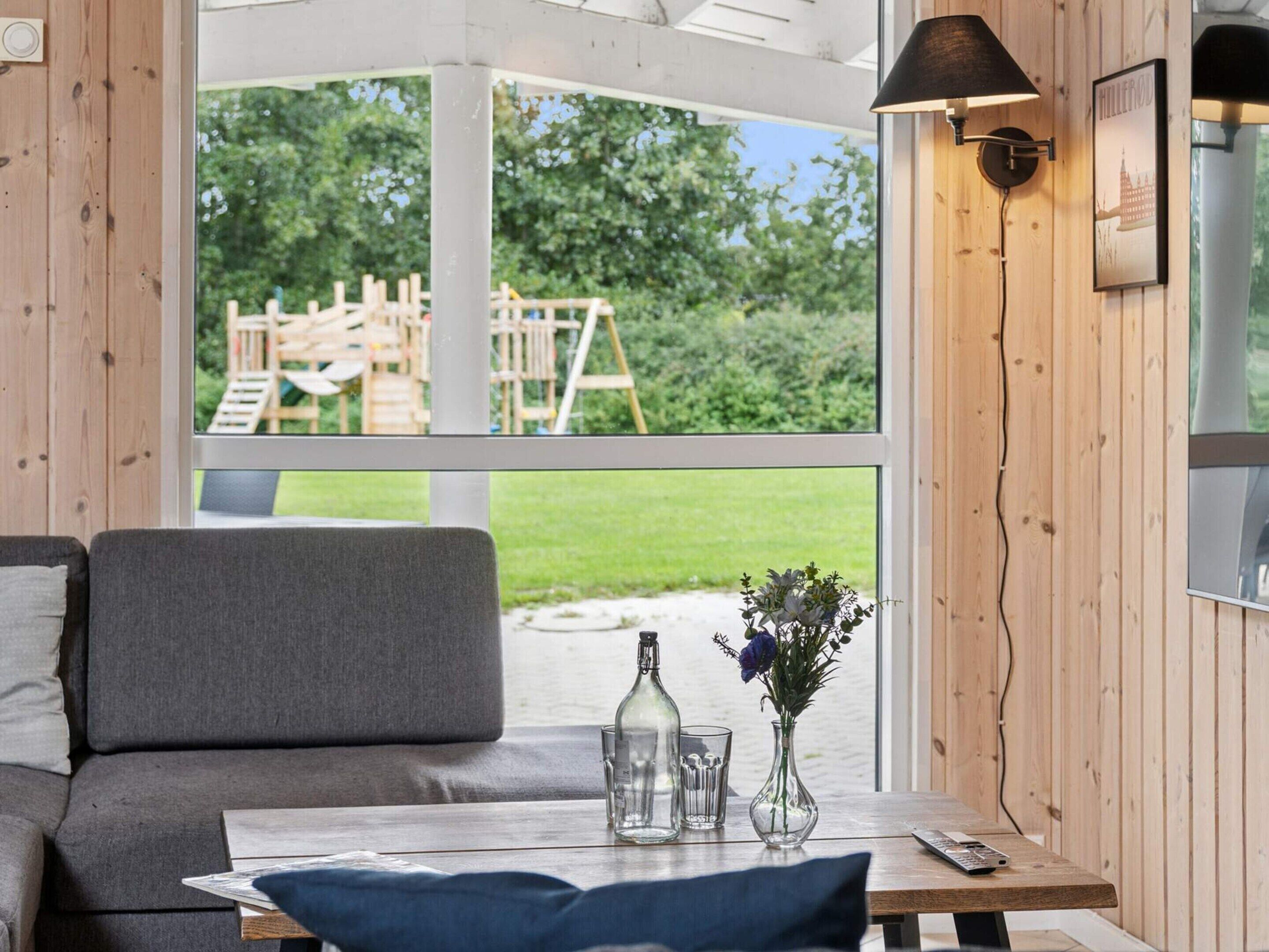 A cozy room with a grey sofa, a wooden table with a vase of flowers, and a view of a wooden deck and greenery outside.