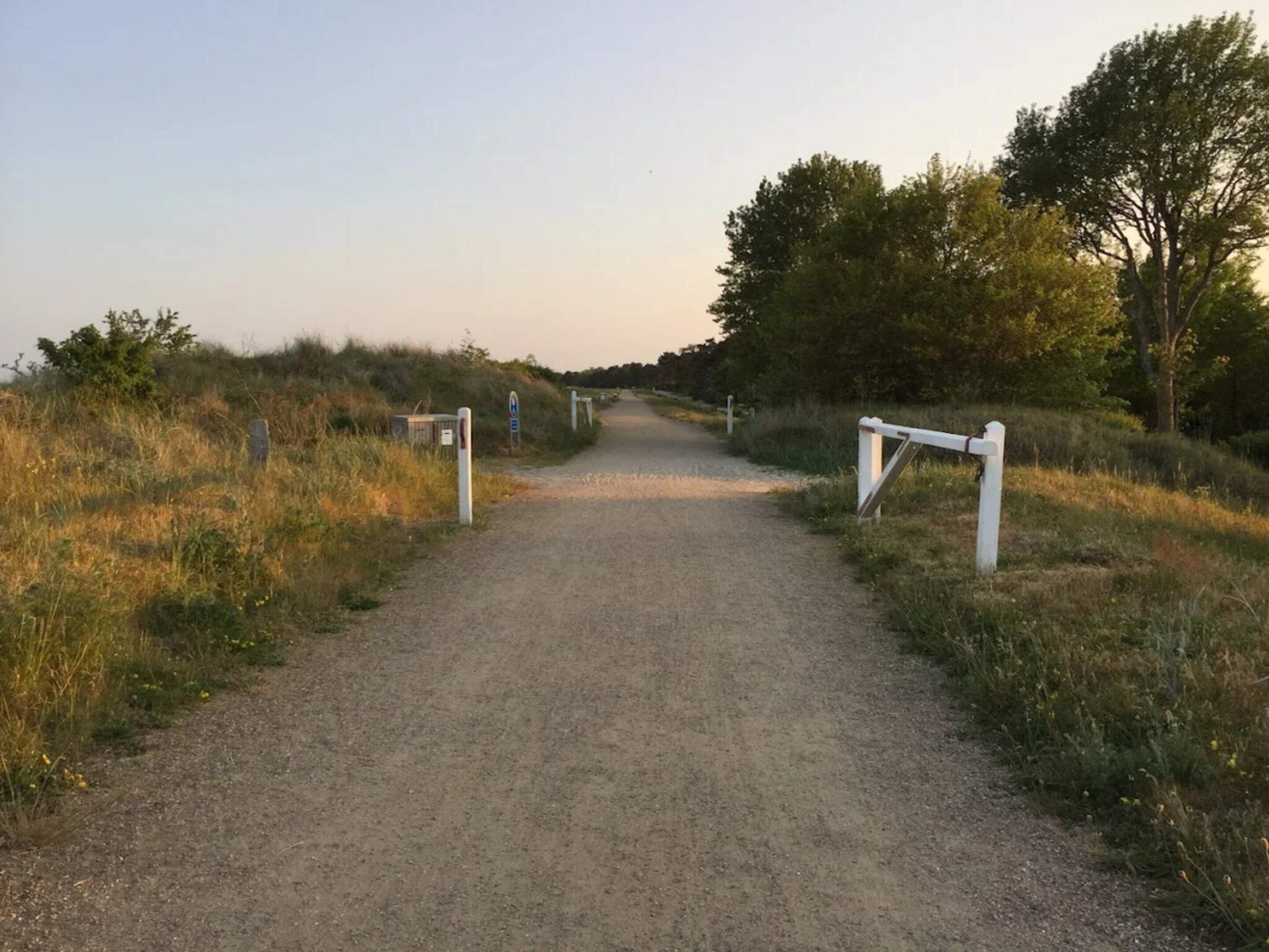 Un chemin de gravier mène à un portail surmonté d’un panneau indicateur, le tout étant entouré d’herbe et d’arbres.