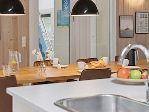 Cozinha moderna com mesa de madeira, cadeiras brancas, pia e uma preparação para o café da manhã com croissants, frutas e café.