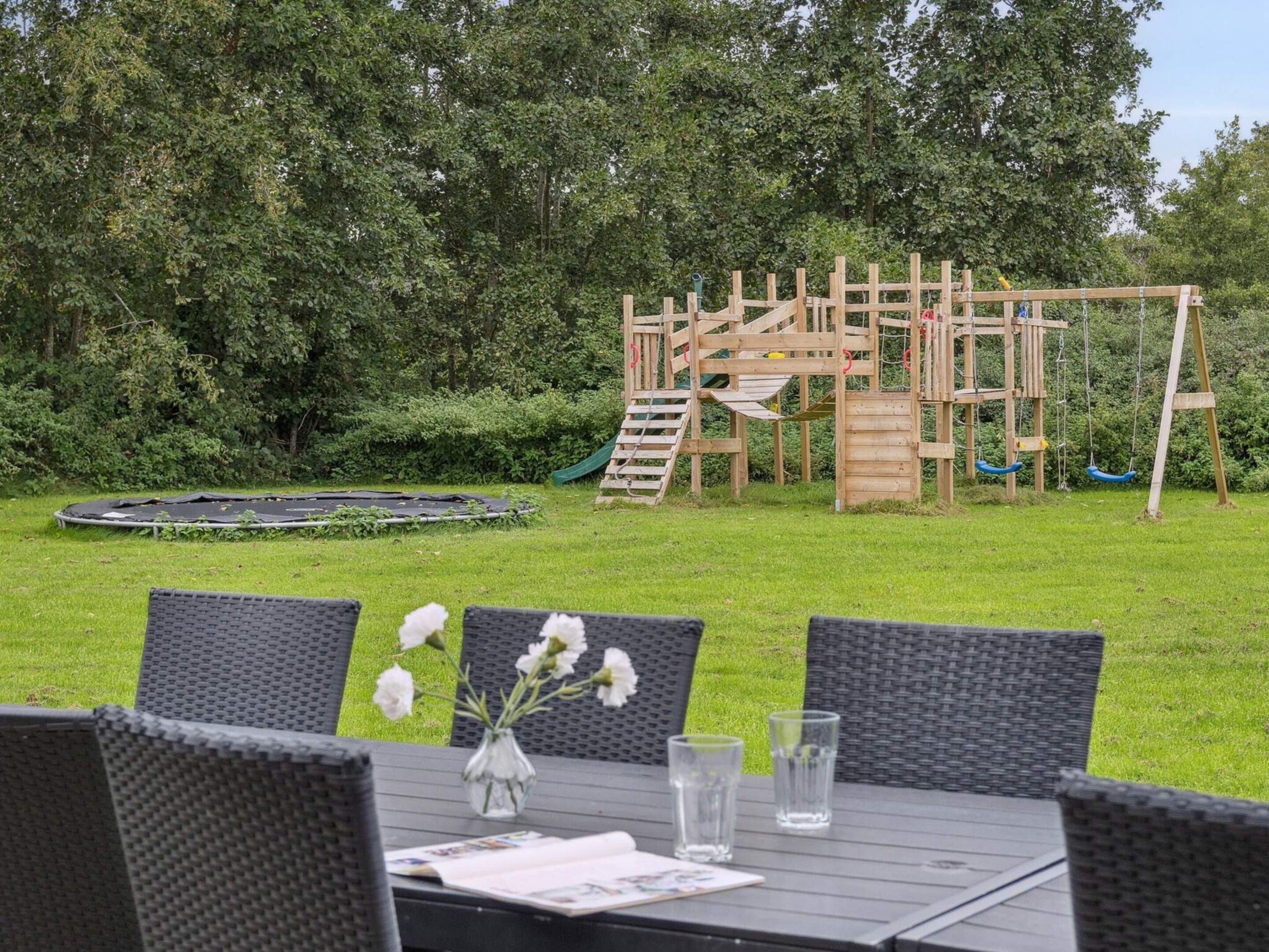A wooden play structure in a grassy area with a trampoline and a table with chairs and a vase of flowers.