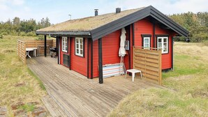 A red wooden cabin with a thatched roof, a wooden deck, and a small table and chair set outside. - 4 Person Holiday Home in Lokken (Lokken)
