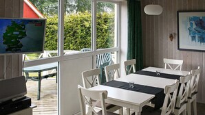 A room with a dining table, chairs, a television, and a window with a view of greenery. - Holiday Home in Gilleleje (Gelting)