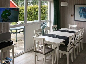 A room with a dining table, chairs, a television, and a window with a view of greenery.