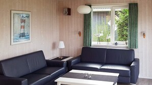 A living room with two black sofas, a white coffee table, a framed painting of a lighthouse, a lamp, and a window with curtains. - Holiday Home in Gilleleje (Gelting)