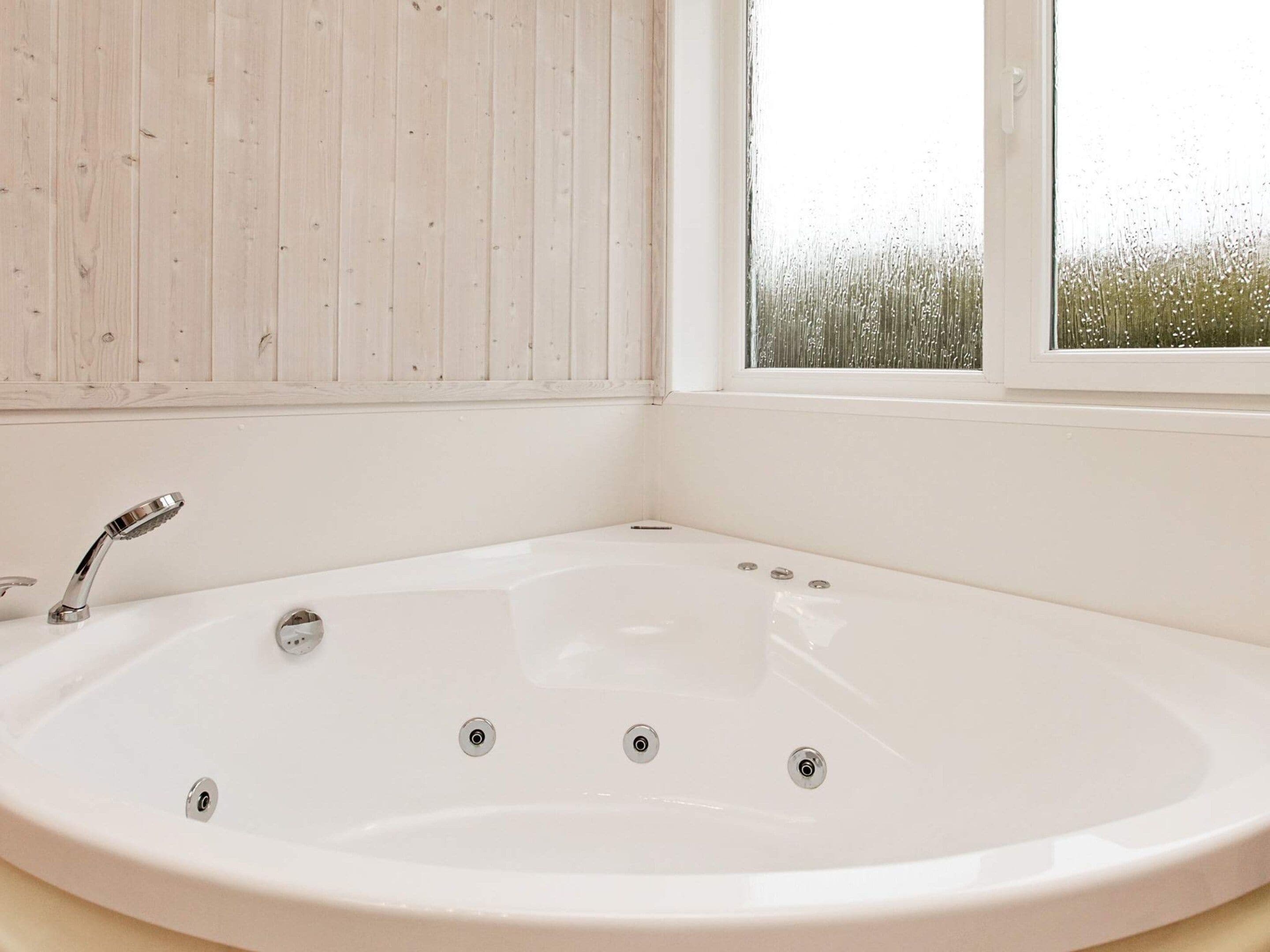 A white bathtub with a modern faucet, a window with a view of greenery, and a wooden wall.