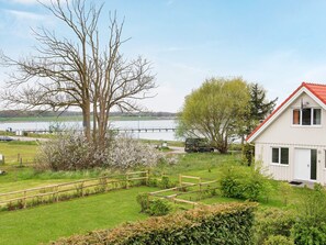 Une maison avec un toit rouge, une clôture en bois et un jardin où poussent différentes sortes de plantes, ainsi qu’un arbre.