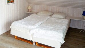 A bedroom with a wooden sloped ceiling, a double bed with white bedding, a bedside table with a lamp, and a wall-mounted painting. - Holiday Home in Gilleleje (Gelting)