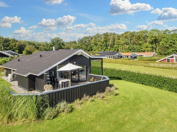 A modern house with a large deck, a dining area with outdoor furniture, and a view of a well-maintained lawn and other houses in the background. - 4 Star Holiday Home in Sjolund (Sjølund)