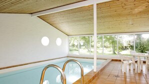 Una piscina interior con paredes de vidrio transparente, suelo de baldosas y una zona de estar techada con mesas y sillas.