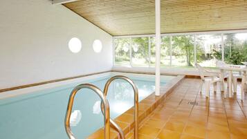 En indendørs swimmingpool med store glasvægge, flisegulv og et overdækket opholdsområde med borde og stole.