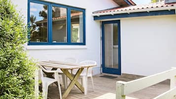 A small, single-story building with a wooden deck, a table with chairs, and a door.