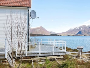 Een houten terras met een witte balustrade, uitzicht op een meer, een schotelantenne op een gebouw en bergen op de achtergrond.