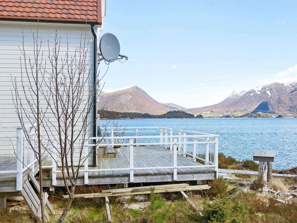 A wooden deck with a white railing overlooking a lake, a satellite dish on a building, and mountains in the background. - Holiday Home in GurskÃ¸y (Heroy)