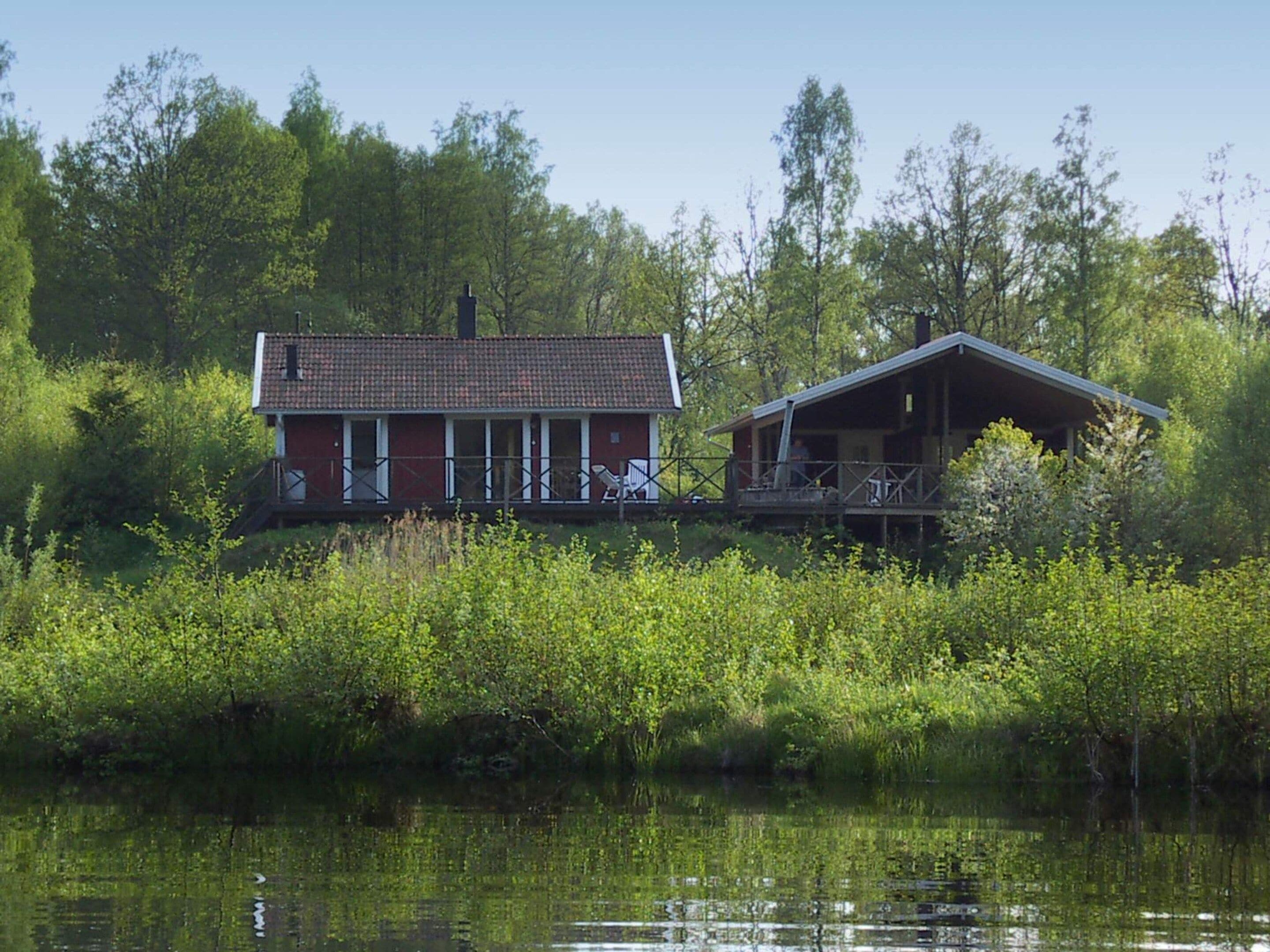 Une cabane dotée d’un porche et d’une terrasse couverte, entourée d’une végétation luxuriante et bordée par un plan d’eau.