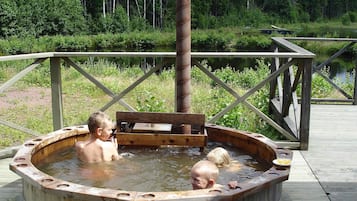Ett utomhusbadkar av trä med en bänk, omgivet av ett trädäck och ett räcke, med ett skogsområde i bakgrunden.