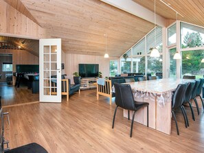 A modern interior with a wooden ceiling, a dining area with a large table and chairs, a seating area with a sofa and TV, and a visible kitchen area.