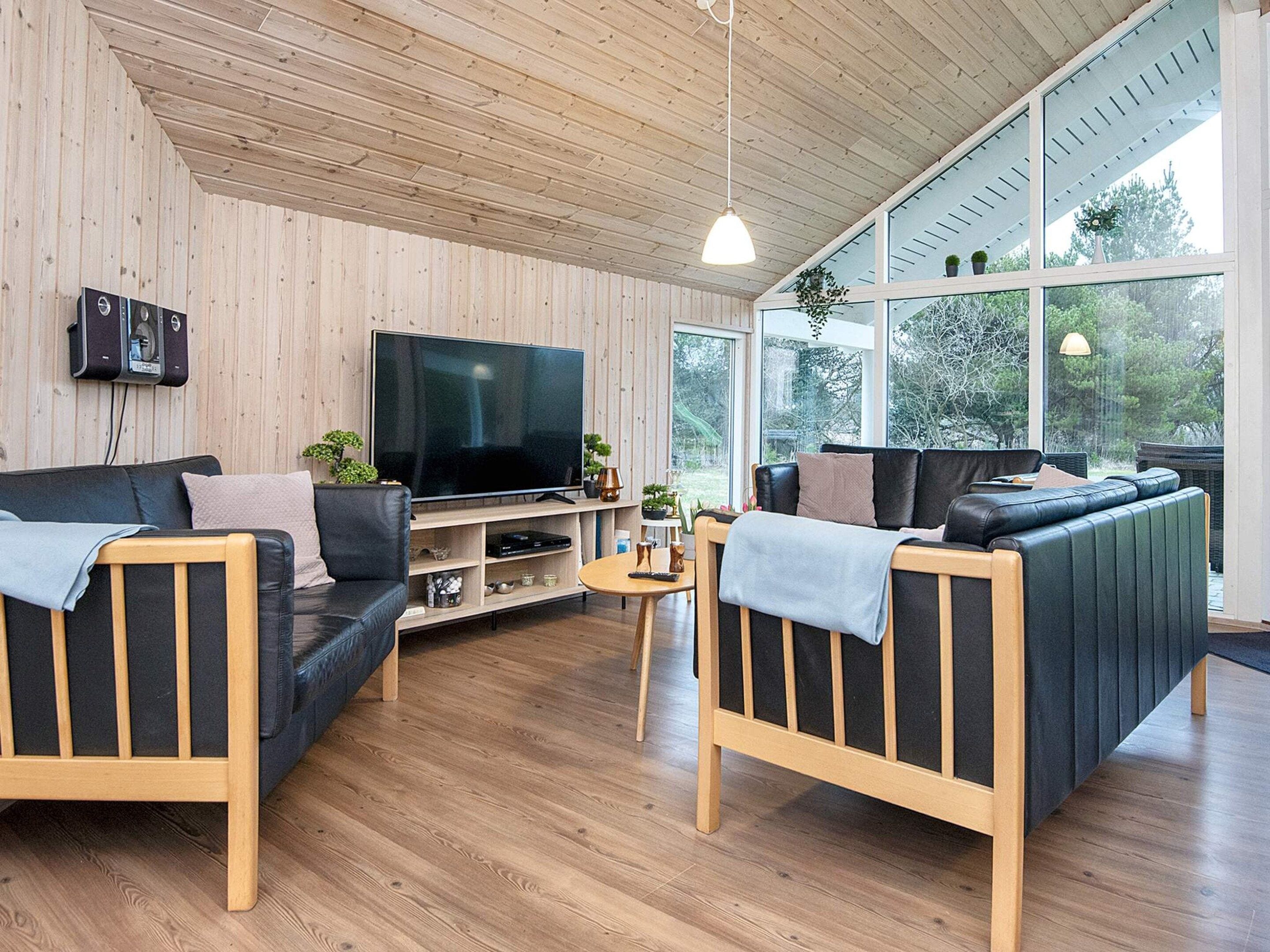 A modern living room with a wooden ceiling, a large flat-screen TV, two sofas, a coffee table, and a large window offering a view of greenery.