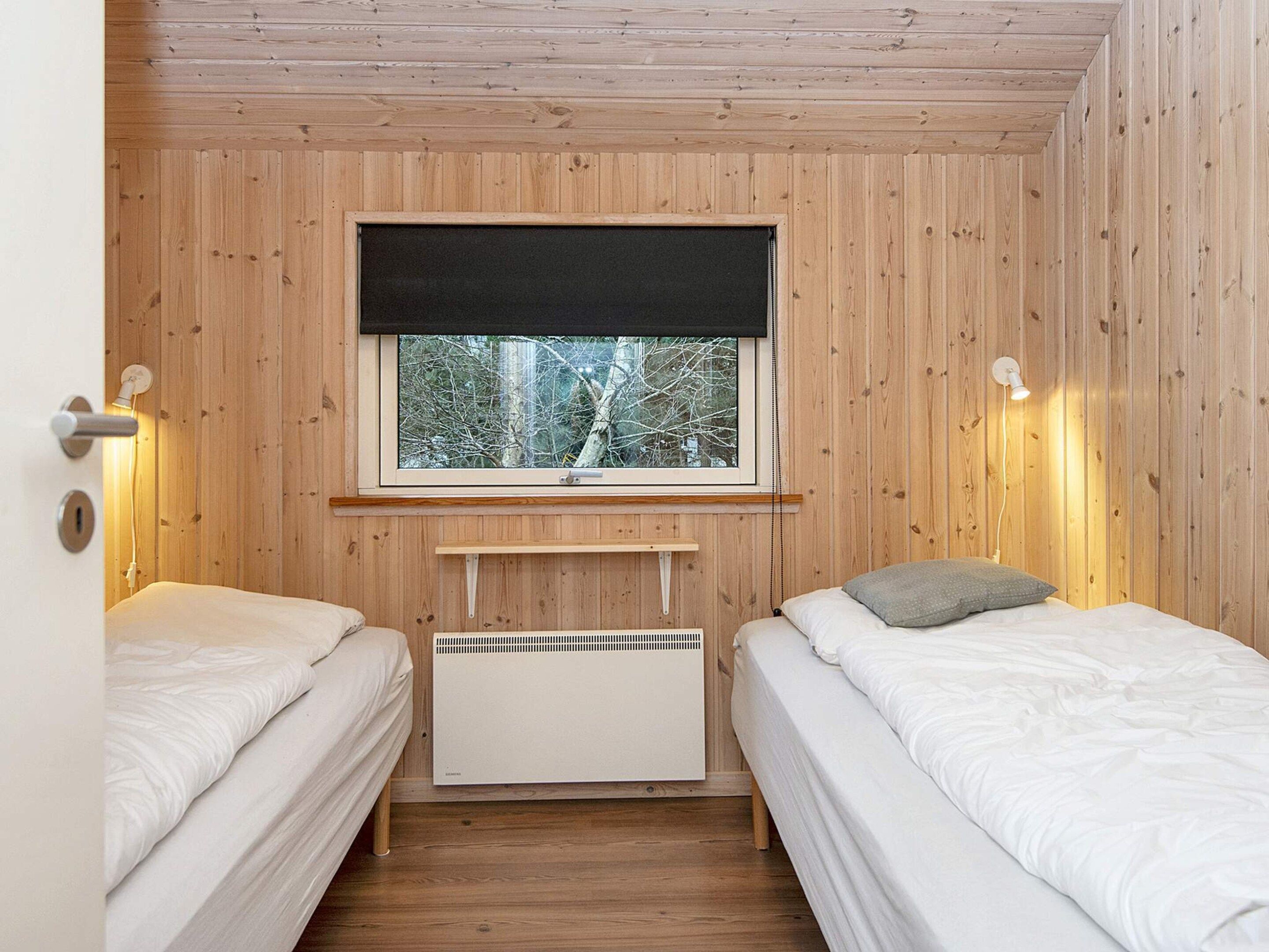A room with two beds, a window with a view of trees, a heater, and a door with a handle.