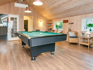 A pool table in a room with wooden walls and ceiling, a wicker chair, and a window with a view of greenery.