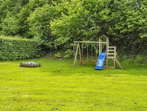 Un parque infantil con tobogán y columpios, rodeado de césped y árboles frondosos.