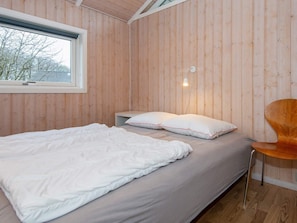 Une chambre avec un mur lambrissé en bois, un lit avec une literie blanche, une table de chevet, une chaise et une fenêtre donnant sur des arbres.