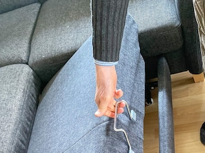 A hand holding a metal object, possibly a tool, near a grey sofa.
