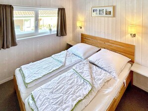 A double bed with a wooden headboard, two bedside lamps, a window with curtains, and a wall-mounted picture frame. - Holiday Home in Otterndorf (Otterndorf)