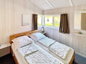 A double bed with white bedding, a bedside table with a lamp, a window with curtains, and a mirror on the wall. - Holiday Home in Otterndorf (Otterndorf)