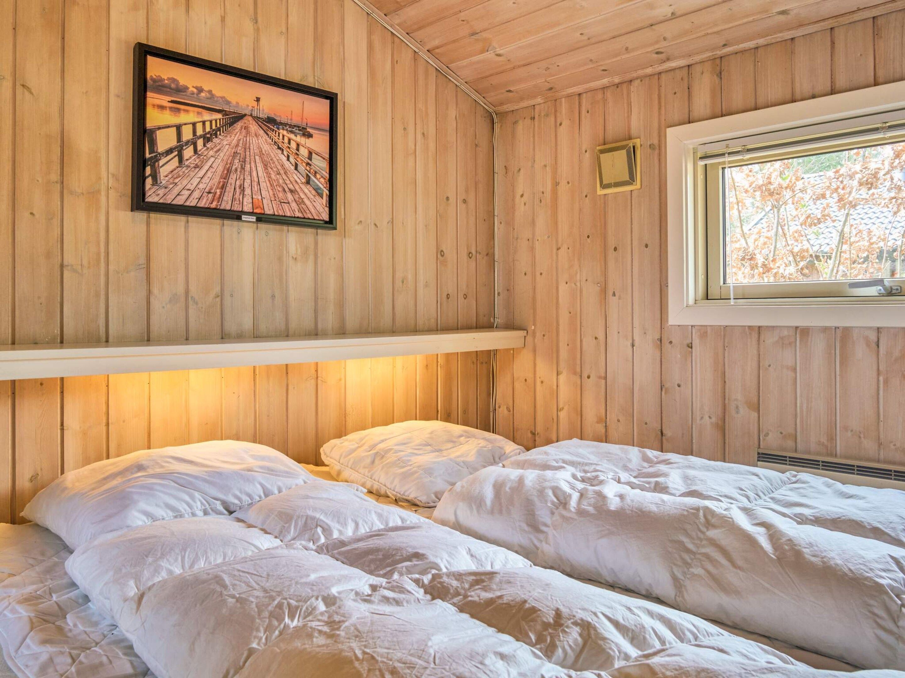 A room with wooden walls, a bed with white bedding, a framed picture of a pier, and a window with a view of trees.