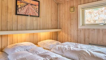 A room with wooden walls, a bed with white bedding, a framed picture of a pier, and a window with a view of trees.