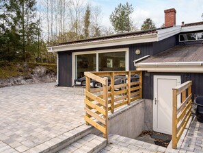 Une maison moderne dotée d’une terrasse en bois, d’un garage et d’une grande fenêtre donnant sur une zone boisée.