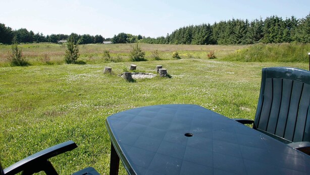 An outdoor seating area with a table and chairs, surrounded by a grassy field and a forest in the background.