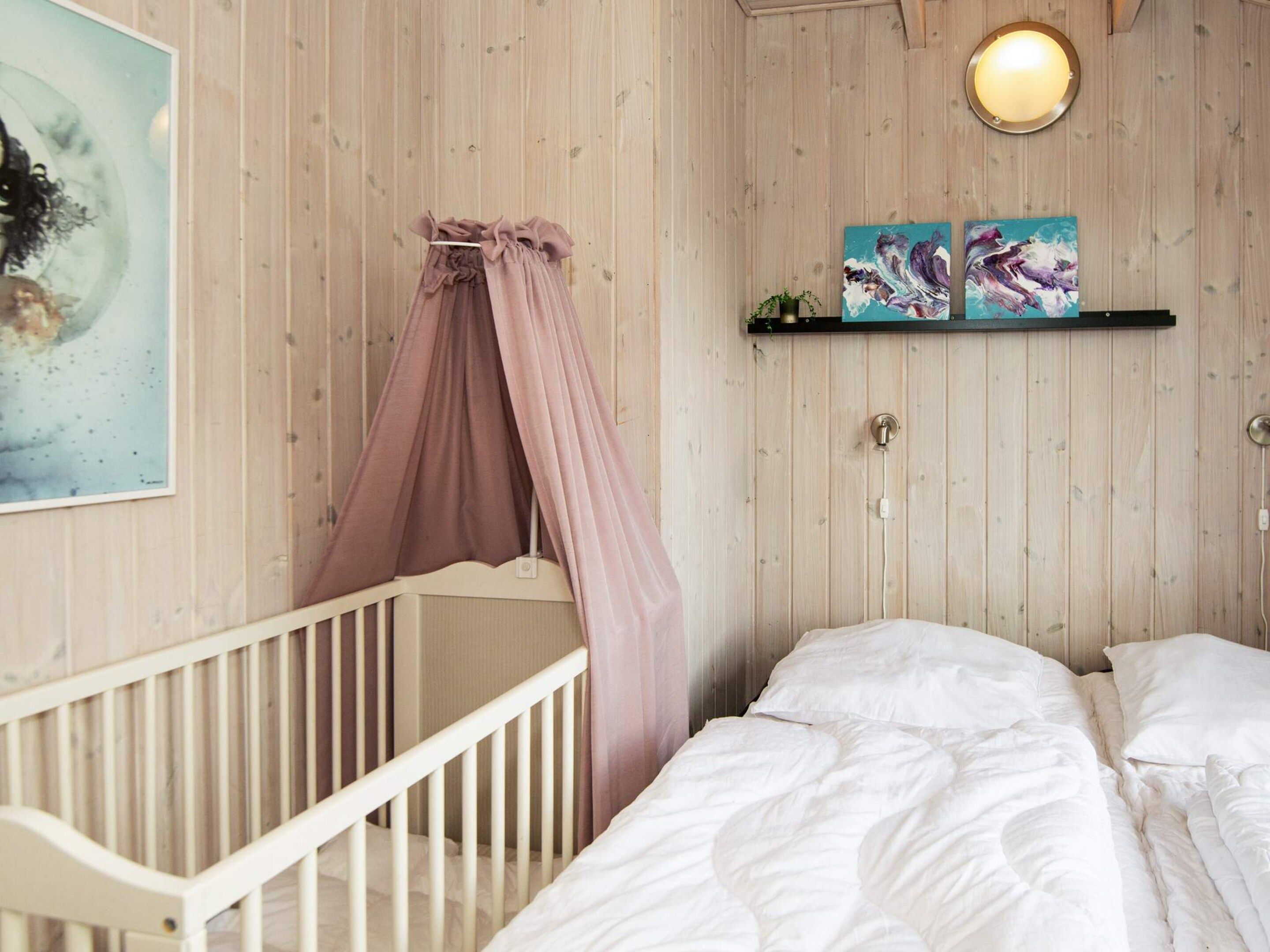 A room with a crib, a bed with white bedding, a wall-mounted shelf with two artworks, and a round wall light.