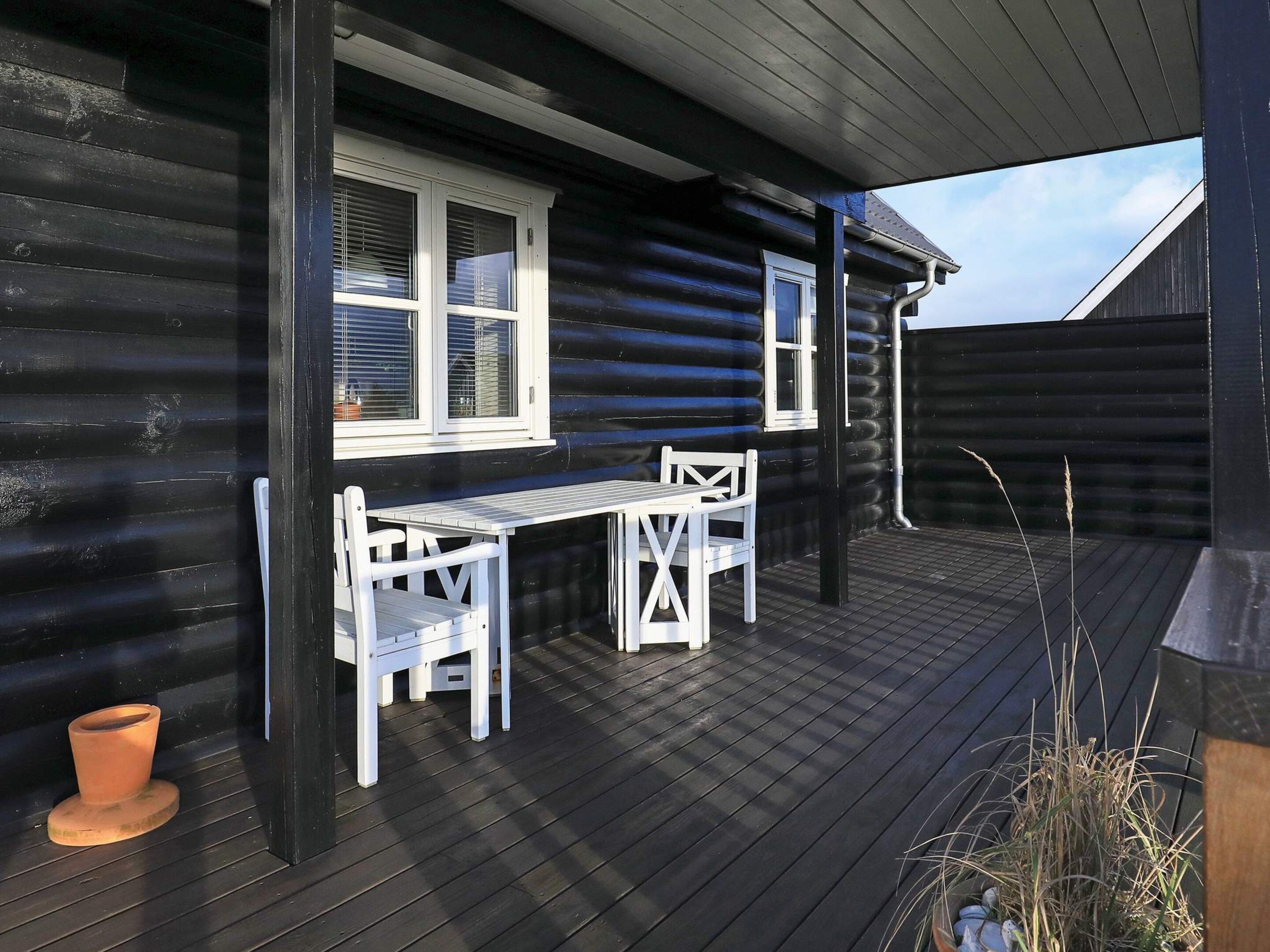 Une terrasse en bois avec une table et des chaises blanches, une fenêtre avec des stores et un petit pot en terre cuite.
