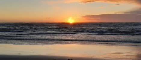 Une plage où le soleil se couche sur l’océan, et où les vagues viennent doucement lécher le rivage.